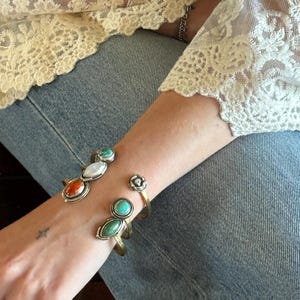 May include: Two metal cuff bracelets worn on the wrist. One bracelet features three oval-shaped stones in turquoise, white, and orange. The second bracelet is a gold-toned open cuff with a small circular floral accent and two green stones.