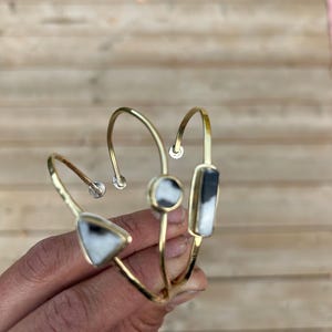May include: Three gold-toned metal cuff bracelets, each featuring a unique stone inlay. One bracelet has a triangular stone, one has a circular stone, and one has a rectangular stone. The stones are white with black marbling and dark grey accents.