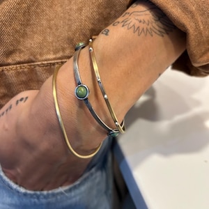 May include: A set of three stacked bangles. One is a plain gold-tone metal band. Another is a silver-tone metal band featuring small raised dots and a single round green stone. The third is a thin gold-tone band with small silver-tone accents.