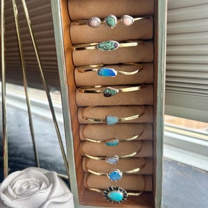 May include: A selection of gold-coloured bracelets, each featuring different gemstones such as turquoise, opal, and pink stones. The bracelets are displayed in a wooden case with a green exterior and a brown interior.