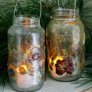 May include: Two decorative glass jar lanterns with silver glitter and dried flower embellishments. Each jar has a wire handle and a lit candle inside, creating a warm glow. The lanterns are decorated with sunflowers, leaves, and other floral elements.