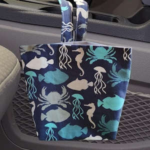 May include: A blue and white fabric car trash bag with a sea life design. The bag has a drawstring closure and is hanging from the headrest of a car seat.