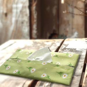 May include: A rectangular fabric tissue box cover in a light sage green color featuring a repeating pattern of white daisy flowers with yellow centers and small white dots. A single white facial tissue is pulled through the center opening.