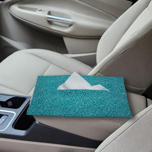 May include: A rectangular teal tissue box cover featuring a white swirling pattern. The cover is made of fabric and has a central opening from which a single white facial tissue is partially pulled out, resting on a beige car center console.