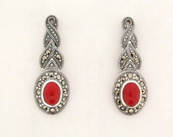 May include: A pair of silver drop earrings. Each earring has a bright red, oval stone set in a detailed silver setting. The top section has a decorative, woven design with small, embedded stones. The earrings are approximately 6 cm long.