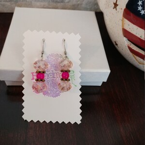 May include: A pair of dangle earrings with pink and fuchsia beads. The earrings feature faceted pink crystal beads and fuchsia-colored round beads, connected by small bronze-colored beads. The earrings are displayed on a white card.