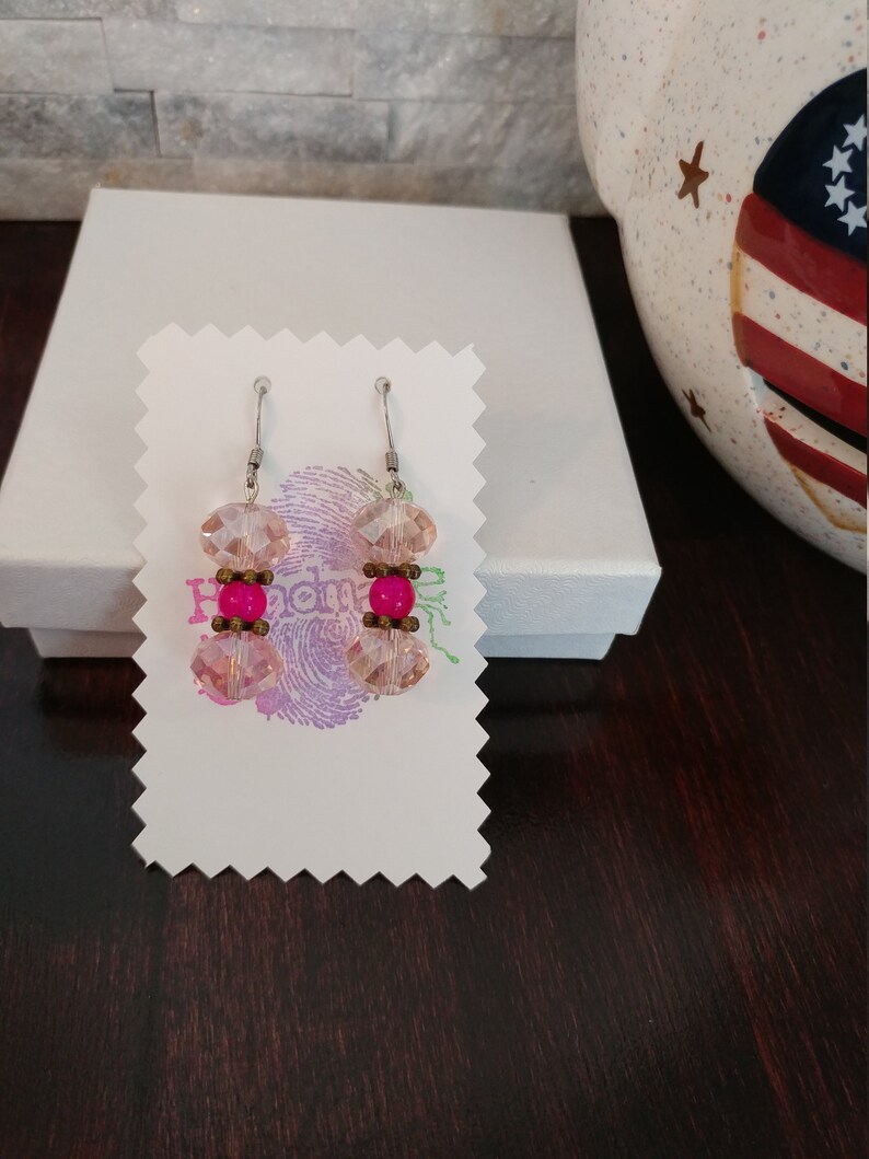 May include: A pair of dangle earrings with pink and fuchsia beads. The earrings feature faceted pink crystal beads and fuchsia-colored round beads, connected by small bronze-colored beads. The earrings are displayed on a white card.