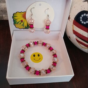 May include: A jewelry set in a white box, featuring a bracelet and earrings. The bracelet and earrings have pink and clear beads with silver hooks. A yellow smiley face sticker is in the box.