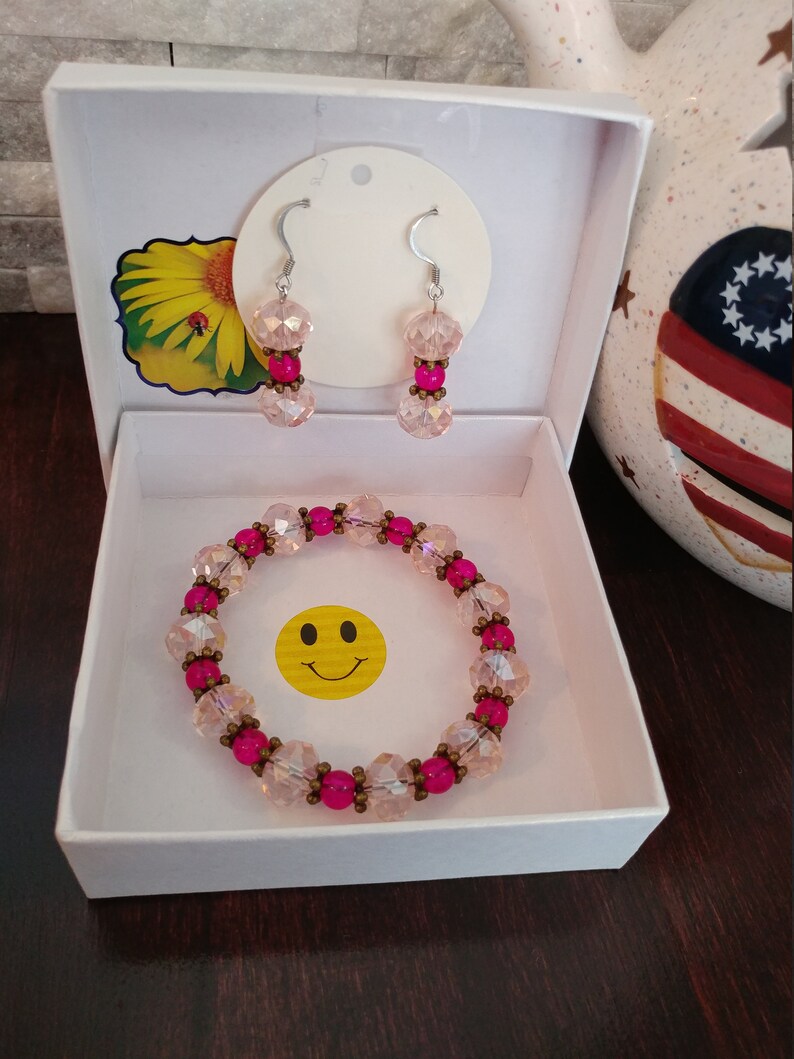 May include: A jewelry set in a white box, featuring a bracelet and earrings. The bracelet and earrings have pink and clear beads with silver hooks. A yellow smiley face sticker is in the box.