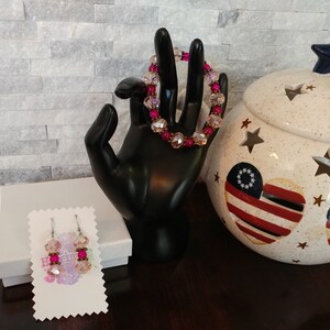 May include: A beaded bracelet and matching earrings with pink and clear beads. The bracelet is displayed on a black hand. The earrings are on a white card. A decorative jar with star cutouts and patriotic heart designs is in the background.
