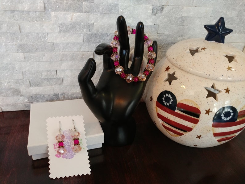May include: A beaded bracelet and matching earrings with pink and clear beads. The bracelet is displayed on a black hand. The earrings are on a white card. A decorative jar with star cutouts and patriotic heart designs is in the background.