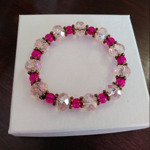 May include: A beaded bracelet featuring alternating pink and clear faceted beads. The bracelet is accented with smaller, round, hot pink beads and antique brass-colored spacers. The bracelet is displayed on a white box.