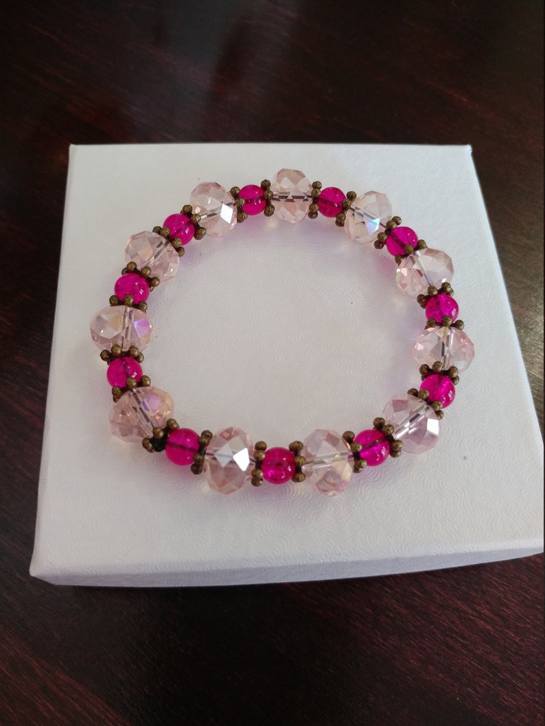 May include: A beaded bracelet featuring alternating pink and clear faceted beads. The bracelet is accented with smaller, round, hot pink beads and antique brass-colored spacers. The bracelet is displayed on a white box.