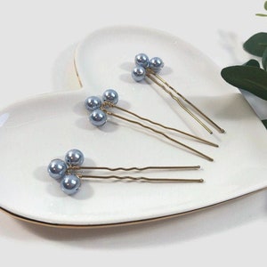 May include: Three hair pins with light blue pearl beads are arranged on a white heart-shaped plate with a gold rim. The pins have a gold-colored metal base. Green leaves are in the background.