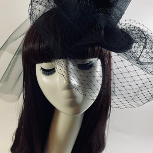 May include: Black top hat with a black veil and netting, labeled "Circus Ringmaster". The hat is on a mannequin head with dark hair and long eyelashes. The veil drapes over the face.