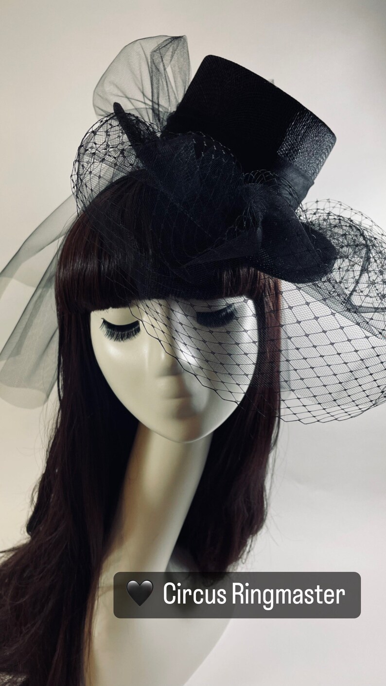 May include: Black top hat with a black veil and netting, labeled "Circus Ringmaster". The hat is on a mannequin head with dark hair and long eyelashes. The veil drapes over the face.