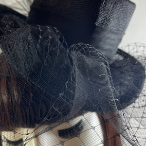 May include: A black hat with a structured crown and a wide brim, adorned with a delicate black netting veil. The hat features a ribbon band and a decorative arrangement of fabric, creating a classic and elegant look. The hat is designed for formal events.