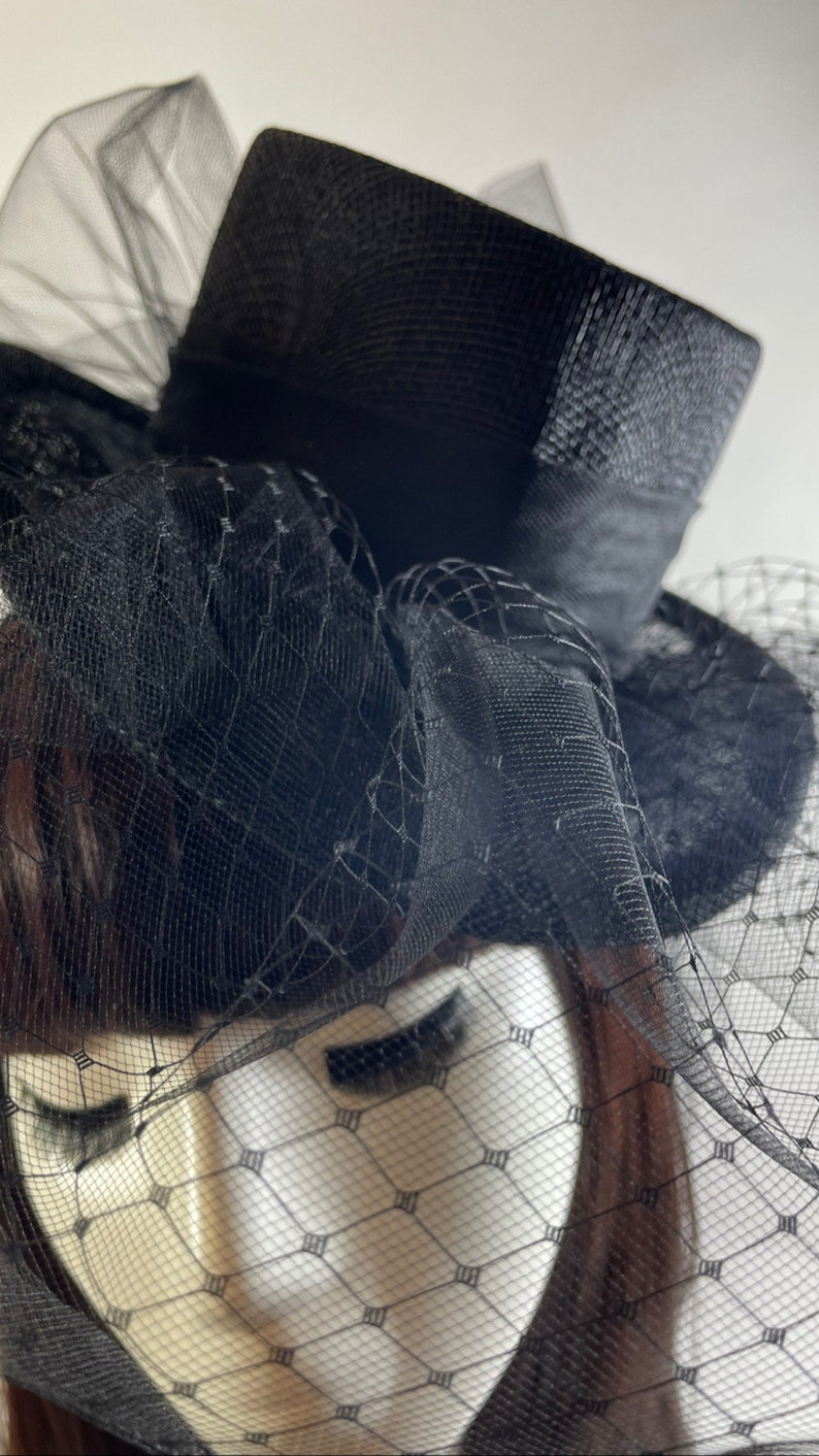 May include: A black hat with a structured crown and a wide brim, adorned with a delicate black netting veil. The hat features a ribbon band and a decorative arrangement of fabric, creating a classic and elegant look. The hat is designed for formal events.