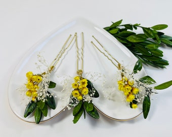 May include: Three gold-toned hair pins, each adorned with a cluster of small yellow flowers, white baby's breath, and green leaves. The pins are arranged on a white, heart-shaped plate, suggesting a floral hair accessory.