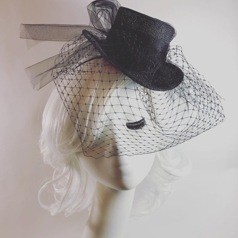 May include: A black hat with a small brim and a sheer veil. The hat is adorned with a decorative bow and is set on a mannequin head with white hair. A single, long eyelash is attached to the veil.