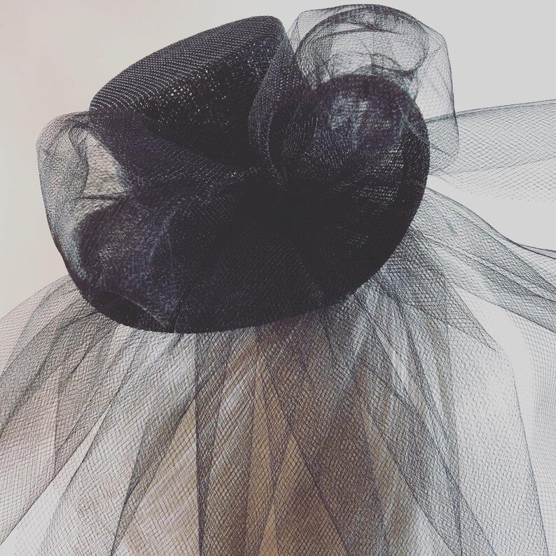 May include: A black fascinator hat with a mesh design. The hat features a rounded base with a large, textured, mesh flower and flowing mesh fabric. The hat is designed to be worn on the head.