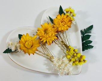 May include: Three yellow chrysanthemum hair pins with gold-colored wire stems and white and yellow accent flowers. The hair accessories are arranged on a heart-shaped white plate with a gold rim, with green fern leaves.
