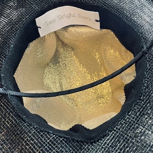 May include: Black mesh hat with a gold-colored interior. The hat features a black ribbon trim and a tag that reads "Two Bright Stars". The hat is designed with a structured shape and a delicate, open weave.