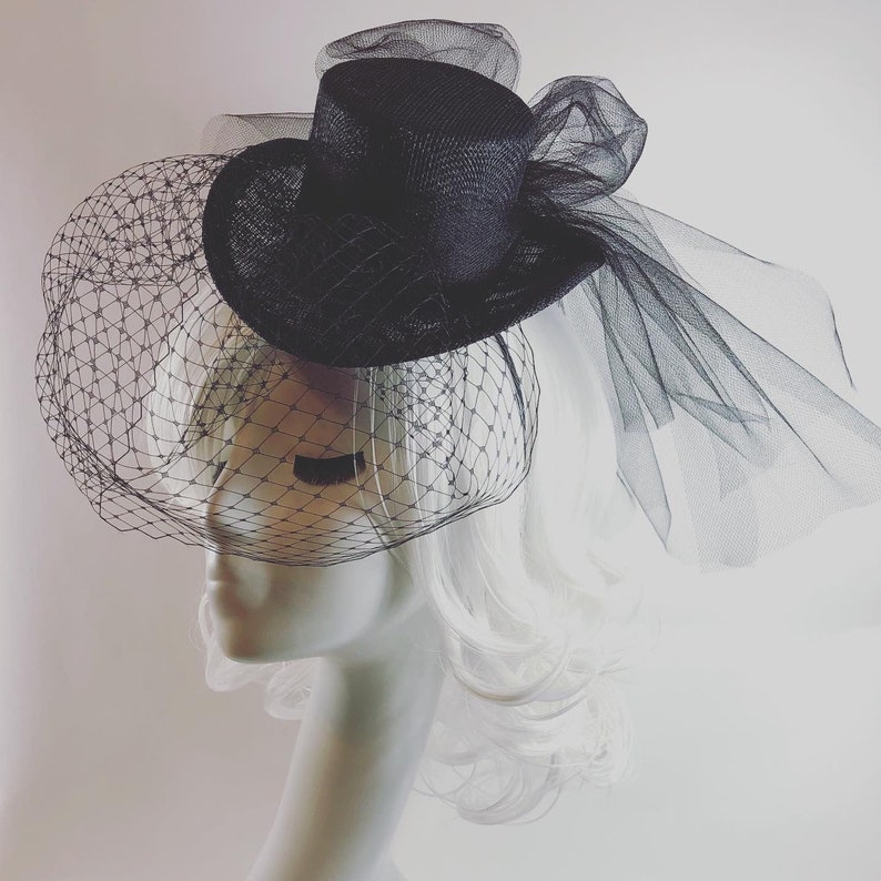 May include: A black hat with a small brim and a tall, cylindrical crown. The hat is adorned with a black veil that drapes over the front and sides. The hat is displayed on a mannequin head with white hair.