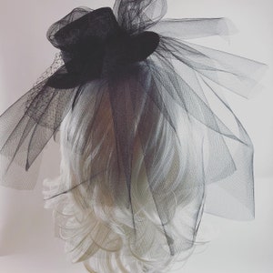 May include: A black fascinator with a small top hat design, adorned with layers of sheer black tulle and a delicate veil. The headpiece is set against a white background, showcasing its elegant and dramatic style. Perfect for special events.