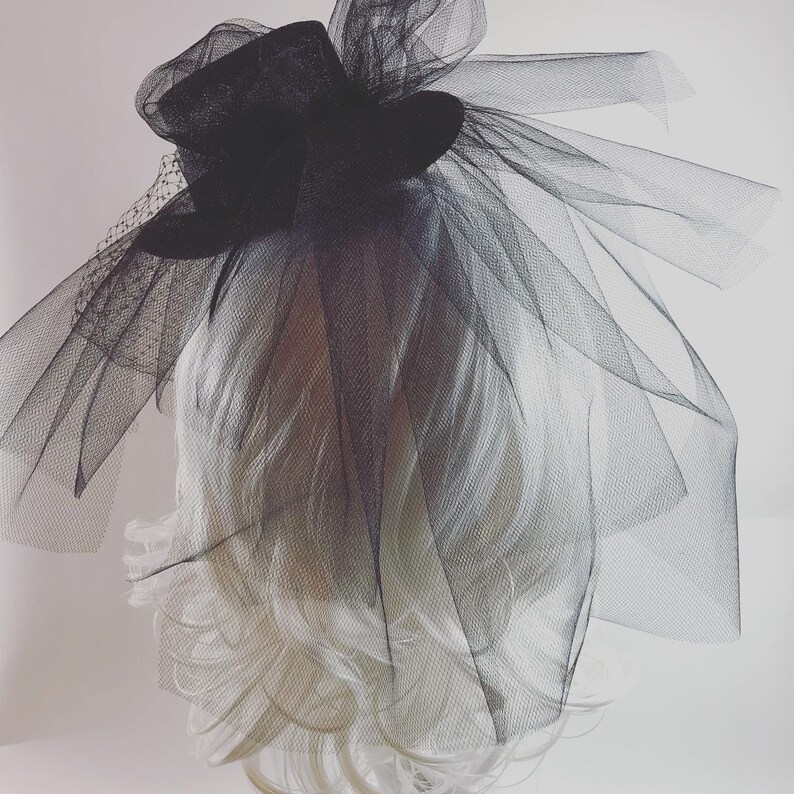 May include: A black fascinator with a small top hat design, adorned with layers of sheer black tulle and a delicate veil. The headpiece is set against a white background, showcasing its elegant and dramatic style. Perfect for special events.