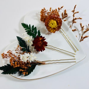 Op de afbeelding: Drie decoratieve haarspelden met gedroogde bloemen. De spelden hebben een verscheidenheid aan bloemen in bruin-, wit- en groentinten. De spelden zijn gemaakt van goudkleurig metaal en zijn gerangschikt op een wit hartvormig bord.