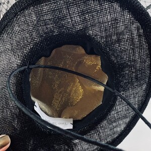 May include: Black hat with a wide brim and a delicate net veil. The hat's interior is lined with gold fabric. A black string is attached to the hat. The hat is likely for special occasions.