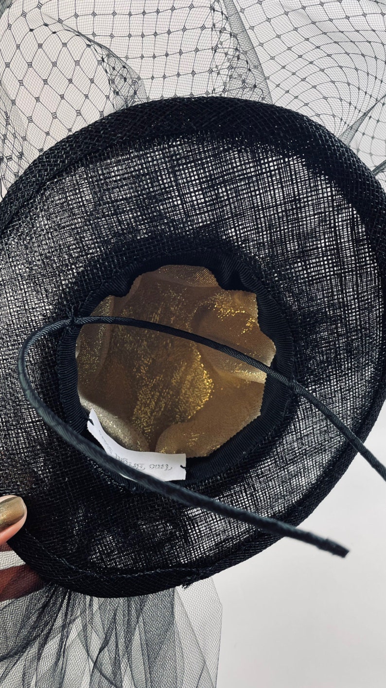 May include: Black hat with a wide brim and a delicate net veil. The hat's interior is lined with gold fabric. A black string is attached to the hat. The hat is likely for special occasions.