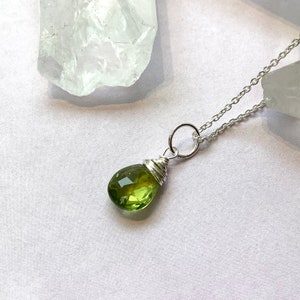 Peut inclure: Un collier en argent avec un pendentif en forme de poire, facetté, en pierre précieuse vert olive. La pierre est enveloppée de fil de fer et suspendue à une chaîne délicate. Le fond est blanc.