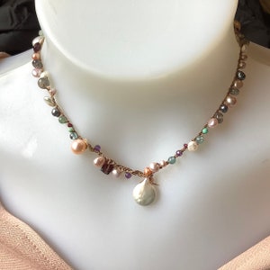 May include: A handcrafted necklace with a woven brown cord, decorated with gemstones and pearls in pink, green, and grey. A large, silver-coloured pendant hangs from the centre.