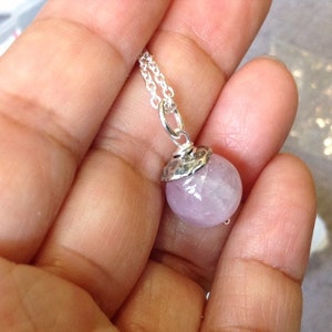 Op de afbeelding: Een zilveren ketting met een ronde, lichtpaarse edelsteen hanger. De gefacetteerde edelsteen heeft een zilveren dop. De ketting is delicaat en zilver. De ketting wordt in een hand gehouden.