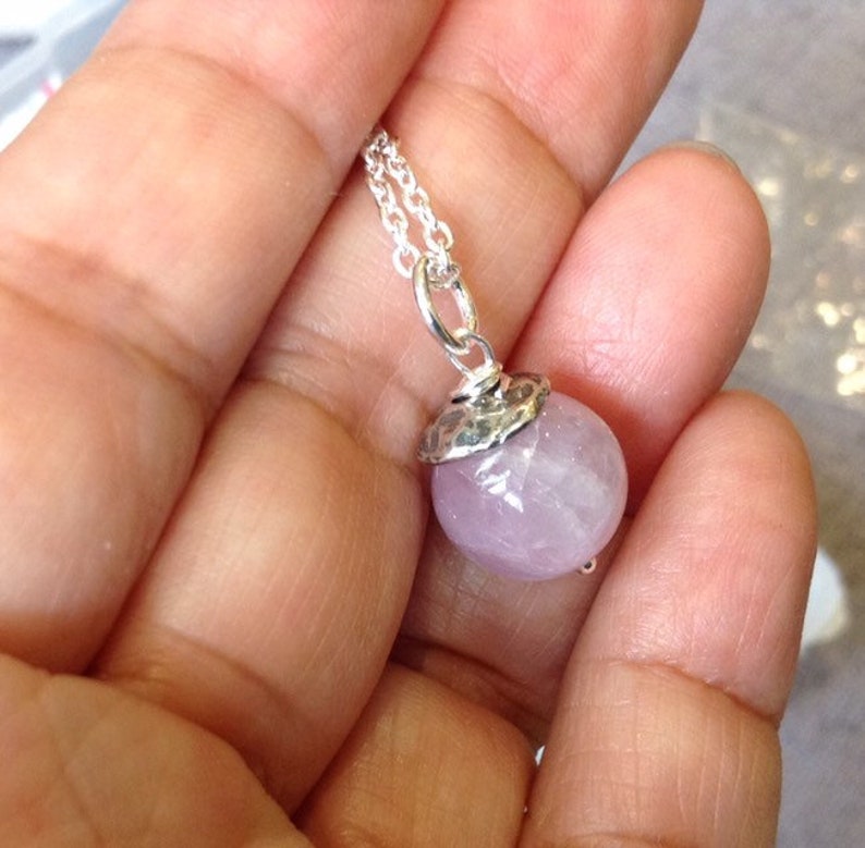 Op de afbeelding: Een zilveren ketting met een ronde, lichtpaarse edelsteen hanger. De gefacetteerde edelsteen heeft een zilveren dop. De ketting is delicaat en zilver. De ketting wordt in een hand gehouden.