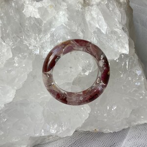May include: A clear resin ring with embedded red and purple stones. The ring is circular and appears to be handmade. The stones are visible throughout the ring, creating a unique and artistic design. The ring is set against a white background.