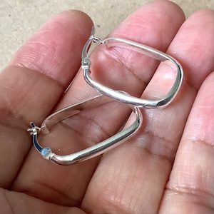 May include: A pair of silver rectangular hoop earrings. The earrings have a smooth, polished finish and a simple, modern design. The hoops are held together by a small clasp.