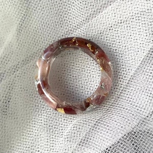 May include: A clear resin ring with embedded red and pink stones and gold leaf accents. The ring has a circular shape and is set against a white, textured background. The ring is a piece of jewelry.