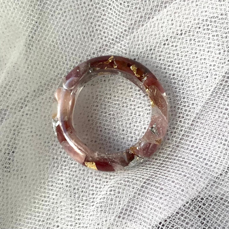May include: A clear resin ring with embedded red and pink stones and gold leaf accents. The ring has a circular shape and is set against a white, textured background. The ring is a piece of jewelry.