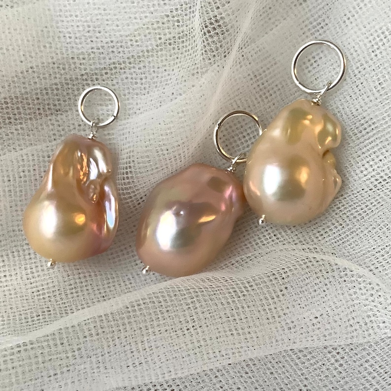 May include: Three baroque pearl pendants with silver-tone findings. The pearls have a lustrous, iridescent sheen with colors ranging from peach to pink. Each pearl is uniquely shaped, with a smooth, irregular surface. The pendants are displayed on a white, textured fabric.