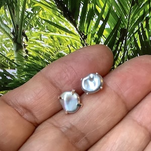 May include: A pair of light grey pearl stud earrings with silver posts. The pearls have an irregular shape and a subtle iridescent sheen. The earrings are displayed against a green and yellow background.