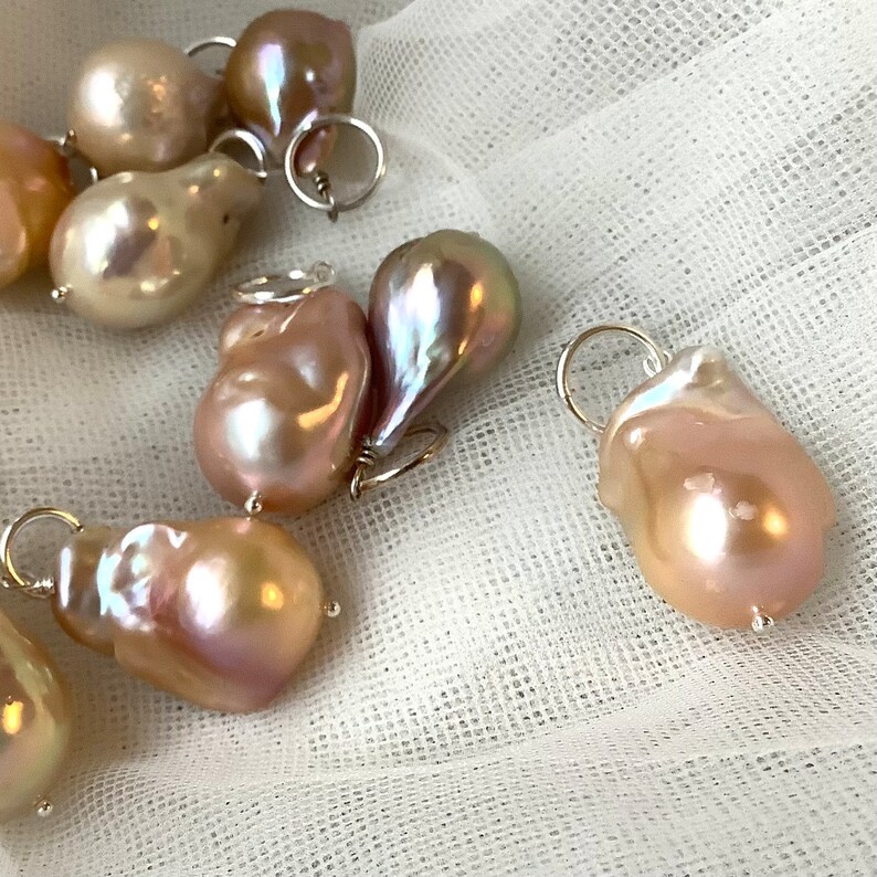 May include: A collection of irregularly shaped, iridescent pearls with silver-tone findings. The pearls display a range of colors, including cream, pink, and lavender, and are set against a white, textured background. These pearls are ideal for jewelry making.