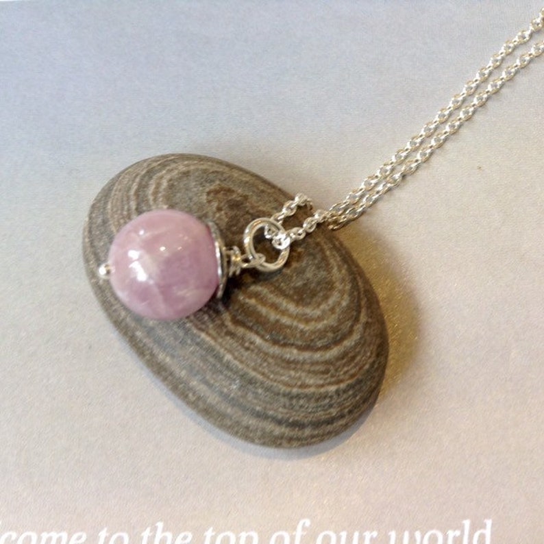 Op de afbeelding: Een zilveren ketting met een ronde, lichtroze edelsteen hanger. De hanger hangt aan een fijne zilveren ketting. De ketting wordt gepresenteerd op een gladde, grijze steen.