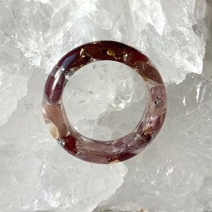 May include: A clear resin ring with burgundy-colored inclusions and gold flakes. The ring is round and sits on a white, textured surface. The inclusions are visible throughout the ring's band.