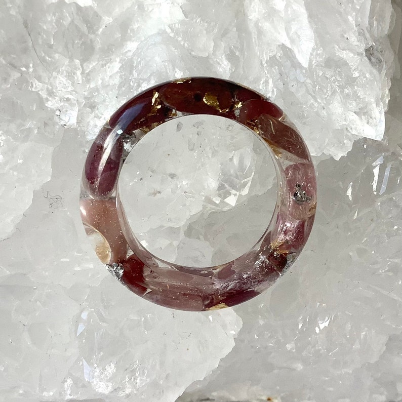 May include: A clear resin ring with burgundy-colored inclusions and gold flakes. The ring is round and sits on a white, textured surface. The inclusions are visible throughout the ring's band.
