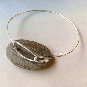 May include: A silver-colored bangle bracelet with an oval-shaped design in the center. The bracelet is resting on a smooth, gray stone. The design is minimalist and modern, suitable for everyday wear.