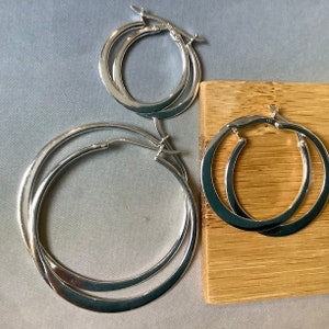 May include: Three pairs of silver hoop earrings in different sizes. The earrings are made of polished metal and have a classic circular design. The hoops are displayed on a light grey surface and a wooden surface.