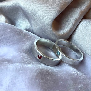 May include: Two silver rings with a hammered texture are displayed on a soft, grey surface. One ring has a red gemstone, the other a blue gemstone. These are likely wedding or engagement rings.